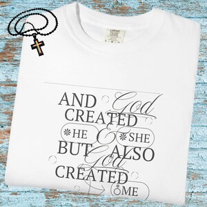 May include: White t-shirt with the text "AND God CREATED *HE *SHE BUT ALSO God CREATED ME". A black beaded necklace with a cross pendant is on the top left. The shirt has a Comfort Colors tag.