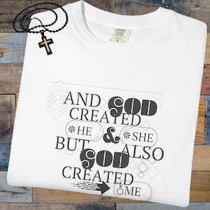 May include: White t-shirt with the text "AND GOD CREATED HE & SHE BUT GOD ALSO CREATED ME" in black. A black beaded necklace with a cross pendant is on the top left. The cross has rainbow colours.