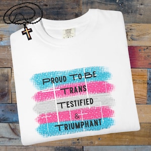 May include: White t-shirt with the text "PROUD TO BE TRANS TESTIFIED & TRIUMPHANT" in black. The text is over a design with blue, pink, and grey brushstrokes. A black beaded necklace with a cross pendant is in the image.