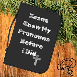 Könnte beinhalten: Schwarzer Bibel-Einband mit dem weißen Text "Jesus Knew My Pronouns Before I Did". Unten befindet sich ein Kreuz-Design mit Fingerabdruck. Ein schwarzer Rosenkranz mit Kreuz liegt auf Holz.