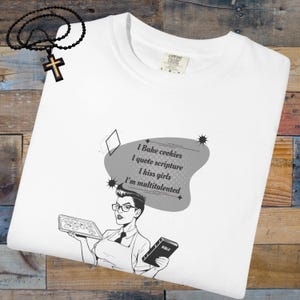 May include: White Comfort Colors t-shirt with a graphic of a woman holding cookies and a Bible. The text reads "I Bake cookies, I quote scripture, I kiss girls, I'm multitalented." A black beaded necklace with a cross is in the upper left.