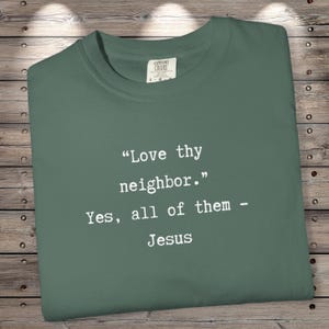 Op de afbeelding: Saliegroen T-shirt met de quote "Heb uw naaste lief. Ja, allemaal - Jezus" in het wit. Het T-shirt heeft een ronde hals en is gemaakt van zacht materiaal. Het label van het merk Comfort Colors is zichtbaar.