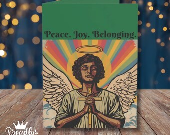Op de afbeelding: Een gevouwen wenskaart met een groen bovengedeelte en de tekst Peace. Joy. Belonging. De illustratie toont een centraal figuur met grote witte vleugels, een aureool en gevouwen handen over een kruis, tegen een achtergrond van een regenboog.