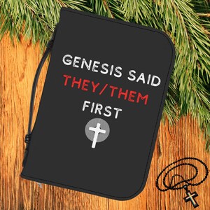 Può includere: Copertina nera della Bibbia con il testo "GENESIS SAID THEY/THEM FIRST" in bianco e rosso. Una croce bianca è centrata sotto il testo. Un rosario nero con una croce è sulla superficie di legno.