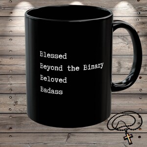 May include: A black ceramic mug with the words "Blessed Beyond the Binary Beloved Badass" printed in white. The mug has a large handle and is set against a wooden background. A beaded necklace with a cross pendant is also visible.