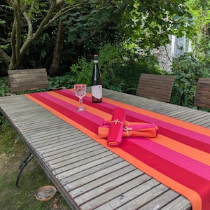 May include: A long rectangular table runner featuring bold horizontal stripes in shades of bright orange, hot pink, and deep red. The runner is placed on a slatted wooden outdoor table with a wine bottle, a glass, and folded napkins tied with twine.