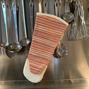 Puede incluir: Un guante de horno a rayas marrón y blanco con un forro de algodón crema. El guante está colgado en una rejilla con varios utensilios de cocina.