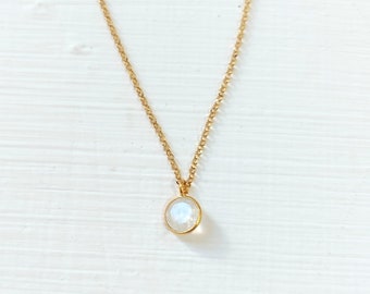 May include: A delicate gold-colored necklace with a small, round, iridescent pendant. The pendant is set in a gold bezel and hangs from a fine chain. The necklace is displayed on a white surface.