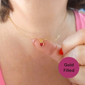 Può includere: Una collana placcata in oro con un piccolo ciondolo a forma di cuore rosso. Il ciondolo a forma di cuore è sospeso a una delicata catena d'oro. L'immagine include un cerchio rosa con le parole "Gold Filled".