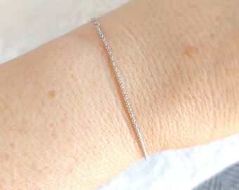 May include: A delicate silver bracelet with a fine chain is shown on a person's arm. The bracelet is simple and elegant, with a subtle sparkle. The background is a soft, neutral color, highlighting the jewelry.