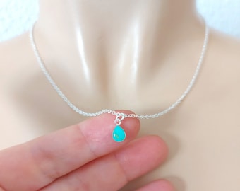 May include: A delicate silver necklace featuring a small, teardrop-shaped turquoise pendant. The pendant is a vibrant teal color, suspended from a fine silver chain. The necklace is displayed on a person's neck, showcasing its simple elegance.