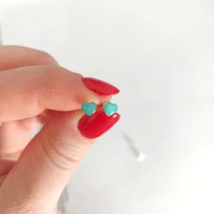 Puede incluir: Un par de pendientes peque&ntilde;os en forma de coraz&oacute;n turquesa. Los pendientes se sostienen entre los dedos con esmalte de u&ntilde;as rojo sobre un fondo blanco.