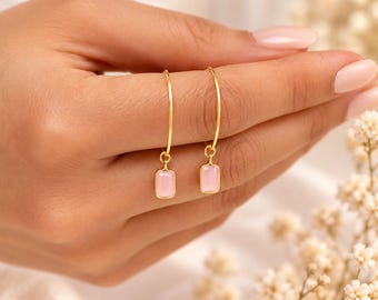 May include: A pair of gold hoop earrings, each featuring a small, rectangular, light pink stone charm suspended from the bottom. The earrings are crafted with a thin gold wire hoop design and a polished metal finish on the stone settings.