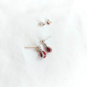 May include: A pair of silver-toned stud earrings featuring a small ball post and a dangling teardrop-shaped charm filled with vibrant red enamel. The set includes two matching earrings and two separate silver-toned earring backs for secure fastening.