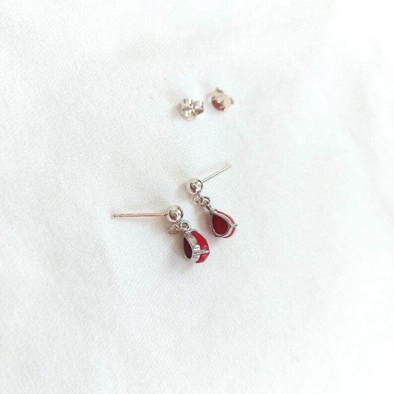 May include: A pair of silver-toned stud earrings featuring a small ball post and a dangling teardrop-shaped charm filled with vibrant red enamel. The set includes two matching earrings and two separate silver-toned earring backs for secure fastening.