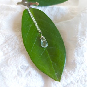 May include: A delicate silver chain necklace with a light blue teardrop-shaped crystal pendant. The pendant is wrapped in silver wire and hangs from a green leaf.