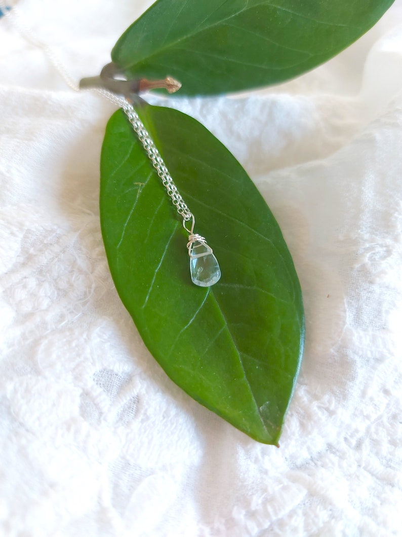 May include: A delicate silver chain necklace with a light blue teardrop-shaped crystal pendant. The pendant is wrapped in silver wire and hangs from a green leaf.