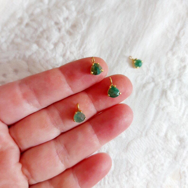 May include: Four small teardrop-shaped green gemstone charms set in gold-toned metal prongs. Each charm features a small circular loop at the top for attachment. The gemstones have a faceted surface and a translucent, deep green color.