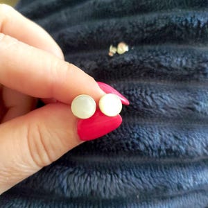 Può includere: Un paio di orecchini a bottone rotondi e bianchi. Gli orecchini sono tenuti tra le dita con smalto rosso brillante, su uno sfondo blu scuro strutturato. Gli orecchini hanno un design semplice e classico.