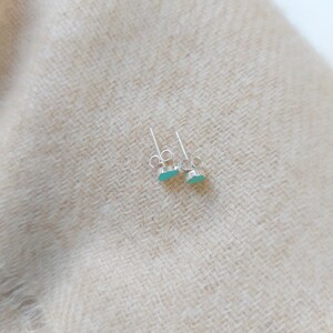 Puede incluir: Un par de peque&ntilde;os pendientes de plata con piedras de color azul claro en forma ovalada. Los pendientes se muestran sobre un fondo de tela beige texturizada. Se ven los postes y los cierres.