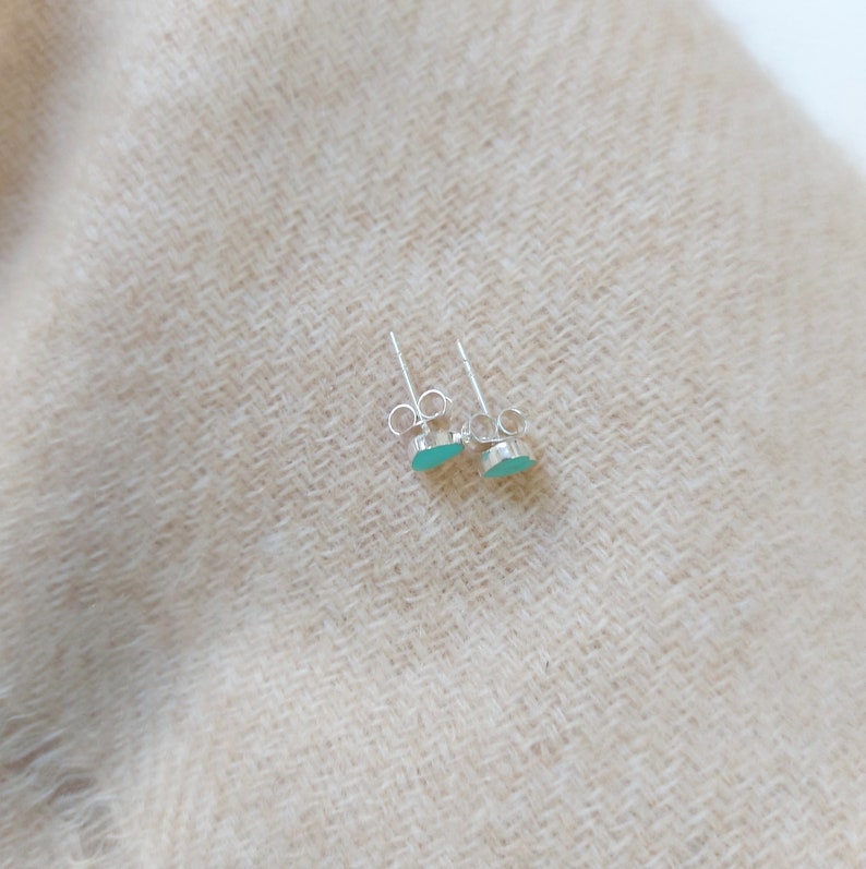 Puede incluir: Un par de peque&ntilde;os pendientes de plata con piedras de color azul claro en forma ovalada. Los pendientes se muestran sobre un fondo de tela beige texturizada. Se ven los postes y los cierres.