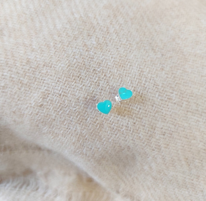 Puede incluir: Un par de pendientes de bot&oacute;n en forma de coraz&oacute;n. Los pendientes tienen un acabado esmaltado turquesa brillante y est&aacute;n montados sobre postes de plata. El fondo es una tela neutra y texturizada, que proporciona un contraste suave.