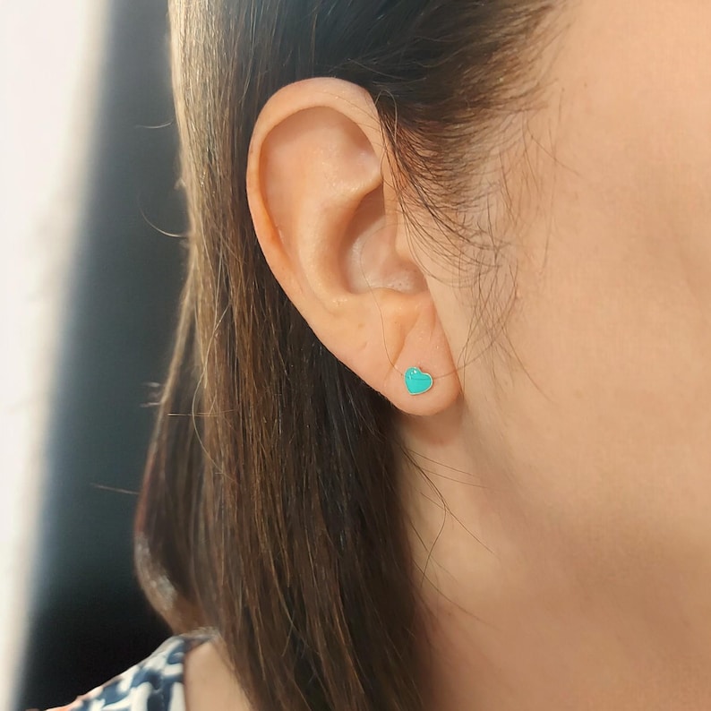 Puede incluir: Primer plano de una oreja con un peque&ntilde;o pendiente de bot&oacute;n en forma de coraz&oacute;n turquesa. El pendiente es de un color vibrante y s&oacute;lido y est&aacute; colocado sobre la piel. El pelo de la persona es casta&ntilde;o y cae detr&aacute;s de la oreja.