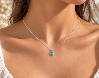 May include: A delicate silver chain necklace featuring a small, teardrop-shaped pendant with a bright turquoise stone. The pendant is securely set in a polished silver frame, hanging centrally from a thin, classic link chain resting on the collarbone.