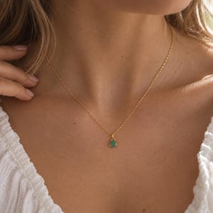 May include: A delicate gold chain necklace featuring a small, triangular-shaped green gemstone pendant. The pendant is securely set in a gold metal frame, suspended from a thin, polished gold link chain that rests against the collarbone area.