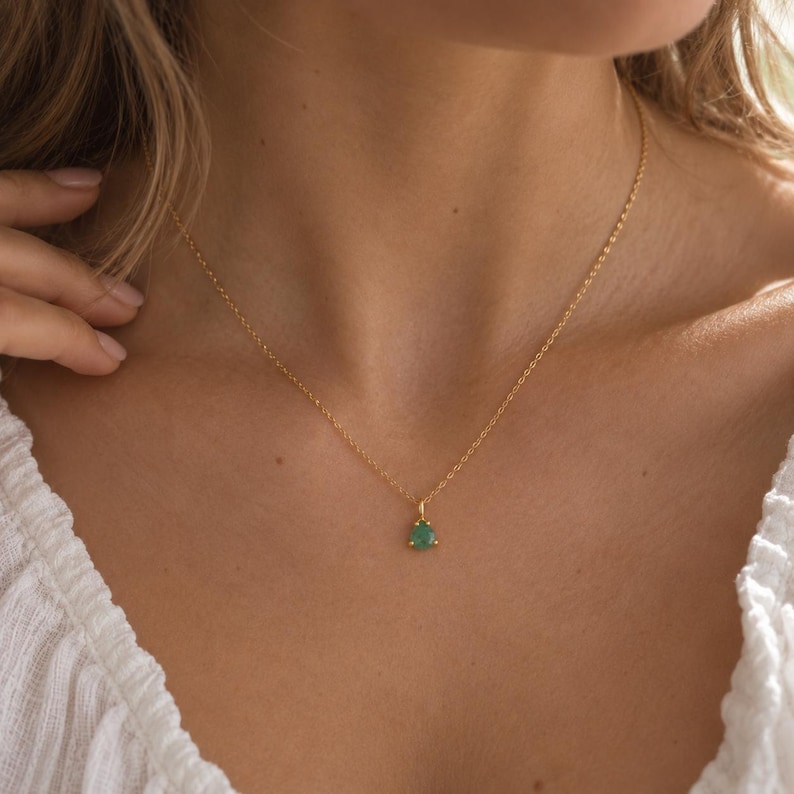 May include: A delicate gold chain necklace featuring a small, triangular-shaped green gemstone pendant. The pendant is securely set in a gold metal frame, suspended from a thin, polished gold link chain that rests against the collarbone area.