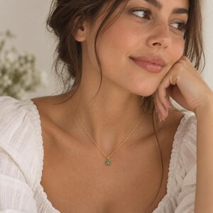 May include: A delicate gold chain necklace featuring a single, small, round-cut green gemstone pendant held in a three-prong setting. The necklace rests against the collarbone, showcasing a thin, twisted metal link design in a bright gold tone.