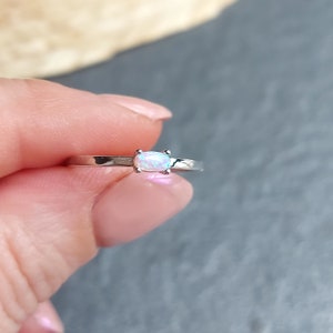Può includere: Un delicato anello d'argento con una piccola pietra di opale ovale. L'opale è di colore bianco latte con sfumature di blu e verde.