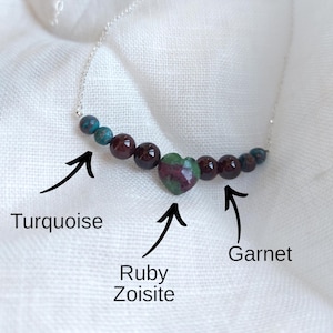 Peut inclure: Un collier délicat avec une chaîne en argent ornée de perles de turquoise et de grenat. Une pierre de rubis zoisite en forme de cœur est le point central. Le collier est présenté sur un fond en tissu blanc.
