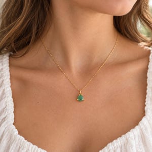 May include: A delicate gold chain necklace featuring a single, faceted, triangular-shaped green gemstone pendant set in a gold bezel. The pendant hangs centrally from a thin, polished gold link chain that rests against the collarbone area.