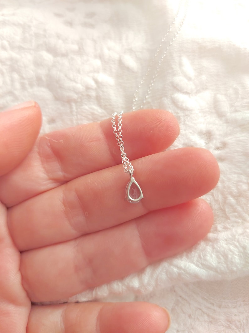 May include: A delicate silver chain necklace with a small teardrop-shaped pendant. The pendant is clear and has a subtle sparkle.