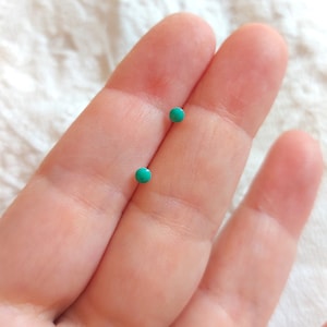 May include: A pair of small, round turquoise stud earrings. The earrings are shown on a person's hand.