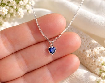May include: A delicate silver chain necklace featuring a small, heart-shaped pendant with a deep blue gemstone set in a smooth silver bezel. The pendant hangs from a simple, thin metal link chain, creating a refined and minimalist jewelry piece.