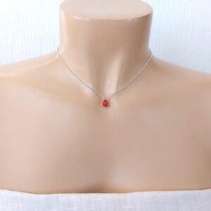 May include: A delicate silver chain necklace featuring a single, small, teardrop-shaped red pendant. The pendant is securely held by a simple metal setting, suspended from a thin, polished silver-tone link chain that rests against the collarbone area.