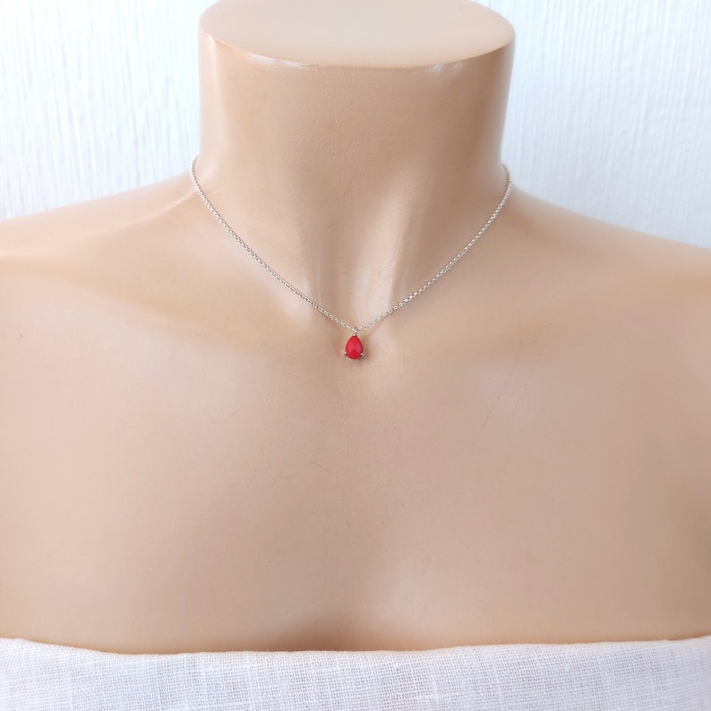 May include: A delicate silver chain necklace featuring a single, small, teardrop-shaped red pendant. The pendant is securely held by a simple metal setting, suspended from a thin, polished silver-tone link chain that rests against the collarbone area.