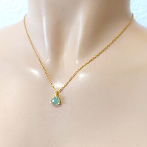 May include: A gold-toned chain necklace featuring a single, small, faceted, light blue-green gemstone pendant. The pendant is secured in a simple gold-toned setting with three prongs, hanging centrally from the delicate, thin metal link chain.