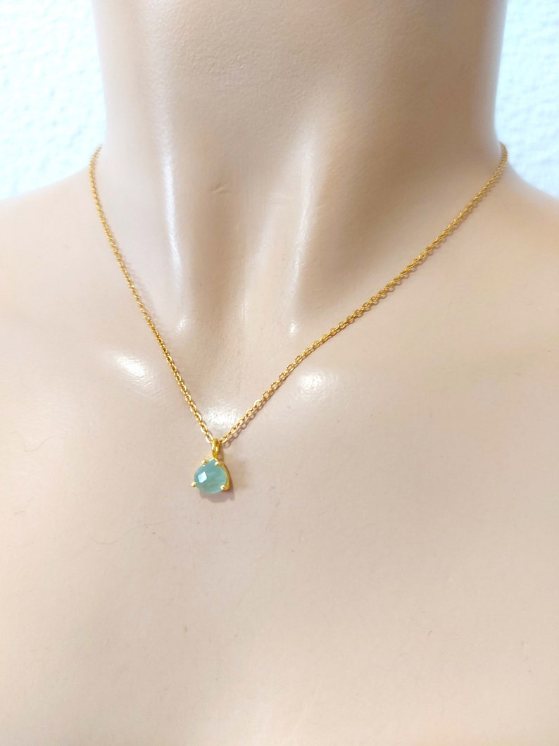 May include: A gold-toned chain necklace featuring a single, small, faceted, light blue-green gemstone pendant. The pendant is secured in a simple gold-toned setting with three prongs, hanging centrally from the delicate, thin metal link chain.