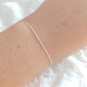 Puede incluir: Un delicado brazalete de cadena de plata con un diseño trenzado. El brazalete se muestra en la muñeca de una persona.