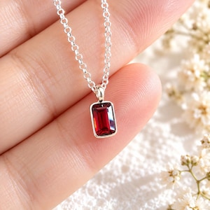 Pode incluir: Um colar de prata com um pingente retangular de pedra preciosa vermelho-escura em uma configuração de metal simples. O pingente pende de uma delicada corrente de prata com elos redondos, exibindo uma superfície facetada que reflete a luz.