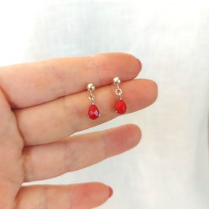 May include: A pair of silver-toned stud earrings featuring small, faceted, teardrop-shaped red stones that dangle from a polished metal ball post. The earrings are designed with a simple drop style, showcasing a vibrant red color and metallic finish.