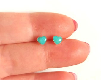 Puede incluir: Un par de pendientes pequeños en forma de corazón. Cada pendiente presenta una piedra de color turquesa engastada en un metal plateado. Los pendientes se muestran en el dedo de una persona.