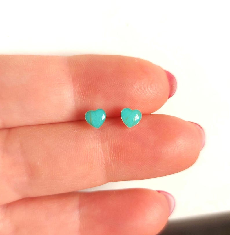 Puede incluir: Un par de pendientes peque&ntilde;os en forma de coraz&oacute;n. Cada pendiente presenta una piedra de color turquesa engastada en un metal plateado. Los pendientes se muestran en el dedo de una persona.