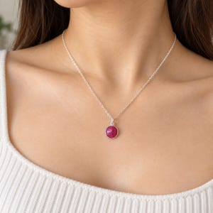 May include: A silver chain necklace featuring a small, round, faceted magenta-coloured gemstone pendant set in a simple silver bezel. The necklace rests against the neck, showcasing the bright pink stone and the delicate metallic link chain design.