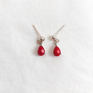 May include: A pair of silver stud earrings featuring small, faceted, teardrop-shaped red stones that dangle from a polished silver ball post. The earrings have a simple, clean design with a metallic finish and vibrant red accents.