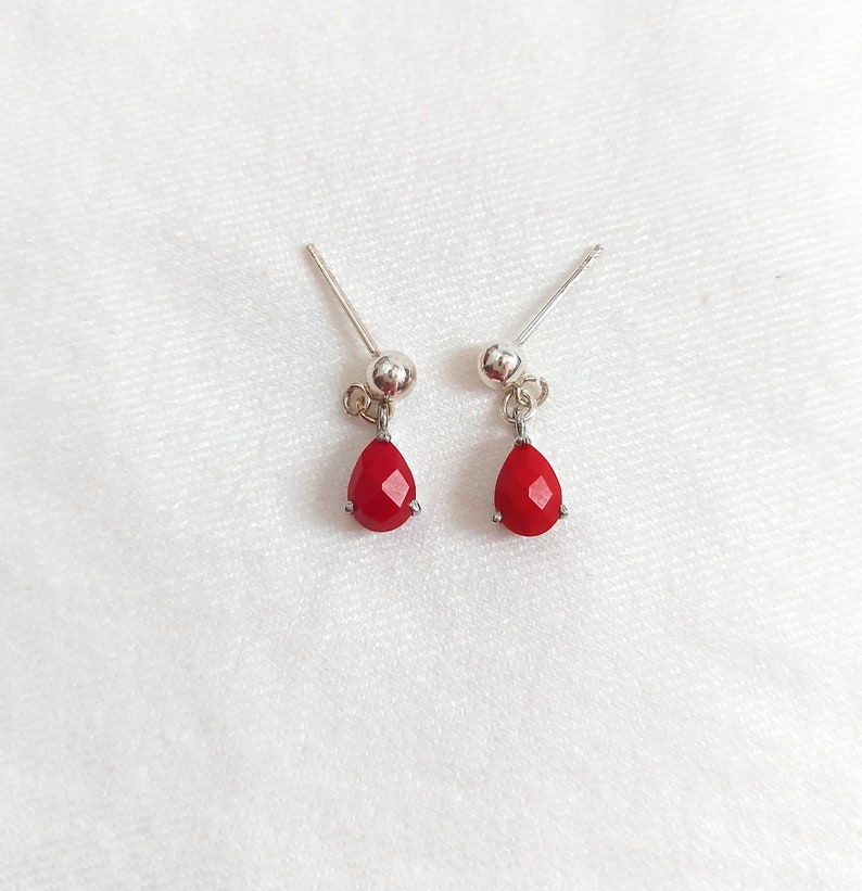 May include: A pair of silver stud earrings featuring small, faceted, teardrop-shaped red stones that dangle from a polished silver ball post. The earrings have a simple, clean design with a metallic finish and vibrant red accents.