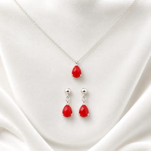 May include: A silver necklace and a pair of matching drop earrings, each featuring a faceted, teardrop-shaped red stone. The necklace has a delicate silver chain, and the earrings are designed with a small silver ball stud and a dangling red pendant.